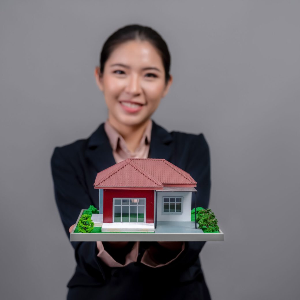 Young Asian woman wearing formal suit holding house model. Enthusiastic Young Asian woman wearing formal suit, holding and showcasing house model on isolated background. Ideal for housing advertisement, home insurance promotions, or customizable copy space. Enthusiastic